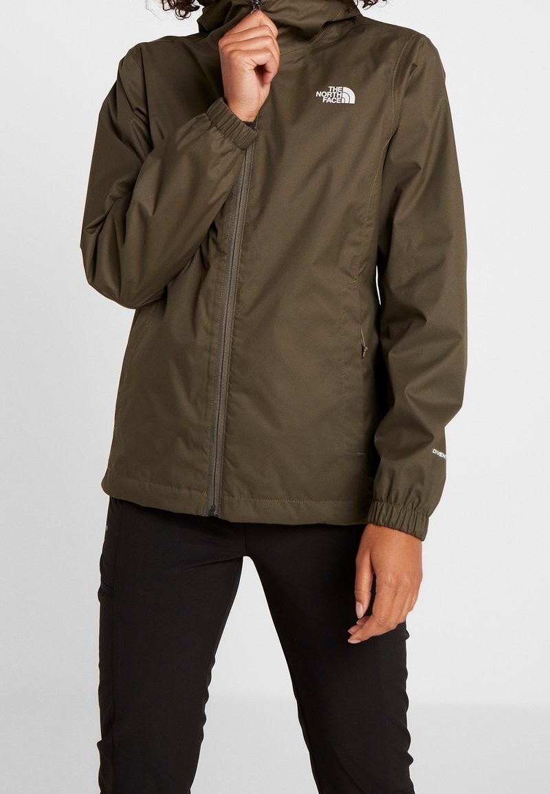 Henkilö, joka käyttää oliivinvihreää vetoketjullista The North Face -takki, jossa on huppu, seisoo yksinkertaista vaaleaa taustaa vasten, käsi kauluksen lähellä.