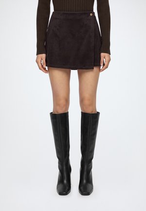 Femme portant une jupe portefeuille marron foncé avec un bouton et des bottes en cuir noires montant jusqu’aux genoux, debout devant un fond uni.