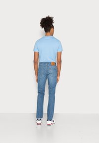 Camiseta de manga corta azul claro, jeans de mezclilla con un parche de cuero marrón y zapatillas blancas con detalles en azul marino, vistas desde atrás.