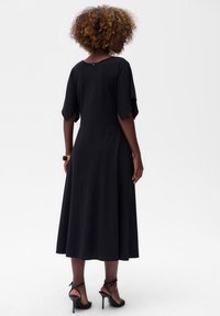 Schwarzes Midi-Kleid aus glattem Stoff mit kurzen, fließenden Ärmeln. Verfügt über ein Schlüssellochdetail im Rücken und einen ausgestellten Saum. Getragen mit schwarzen, riemenartigen Absätzen.