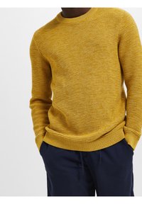 Gelber Strickpullover mit strukturierter Oberfläche, geripptem Rundhalsausschnitt und engen Bündchen. Kombiniert mit dunkelblauen Hosen mit elastischem Bund.