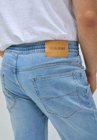 Jeans de mezclilla azul claro con una textura suave, que presentan una etiqueta de cuero marrón claro marcada "SALSA JEANS" y dos bolsillos traseros con detalles de costura.