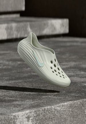 Jasnozielony but wsuwany z otworami wentylacyjnymi i turkusowym logo Nike, unoszący się nad kamiennym podłożem z schodami w tle.