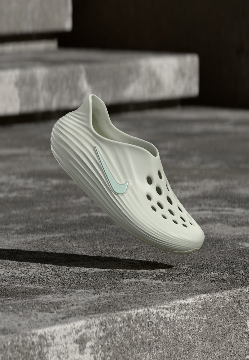 Šviesiai žalia batai be raištelių su ventiliacijos skylutėmis ir turkio spalvos „Nike“ logotipu, plūduriuojantys virš akmeninės dangos, kurioje fone matosi laiptai.