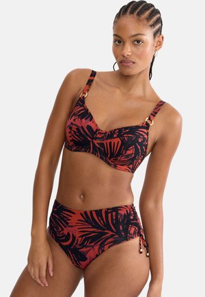 Kvinde med flettet hår iført en rød og sort bikini med tropisk print, højtaljede underdele og justerbare stropper, stående mod en ensfarvet baggrund.