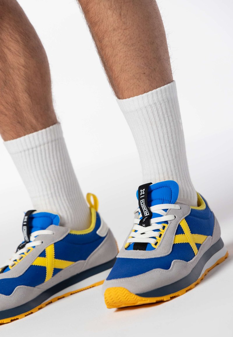 Baskets bleues et grises avec des accents jaunes, présentant un dessus en tissu texturé, des lacets blancs et une semelle en caoutchouc à motifs. Des chaussettes blanches côtelées sont visibles.