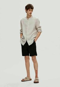 Light gray linen button-up shirt, black shorts, and tan sandals; relaxed fit; rolled sleeves; visible tattoo on the leg.