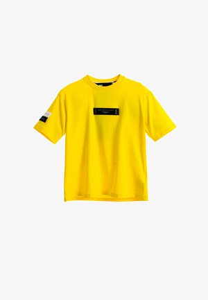 Camiseta amarilla de algodón con mangas cortas, con una etiqueta rectangular negra en la parte delantera y dos etiquetas de acento en las mangas.