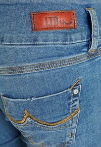 Jeans de mezclilla azul con una etiqueta de cuero marrón de LTB, costuras amarillas en contraste y un pequeño bolsillo trasero en ángulo con un remache metálico.