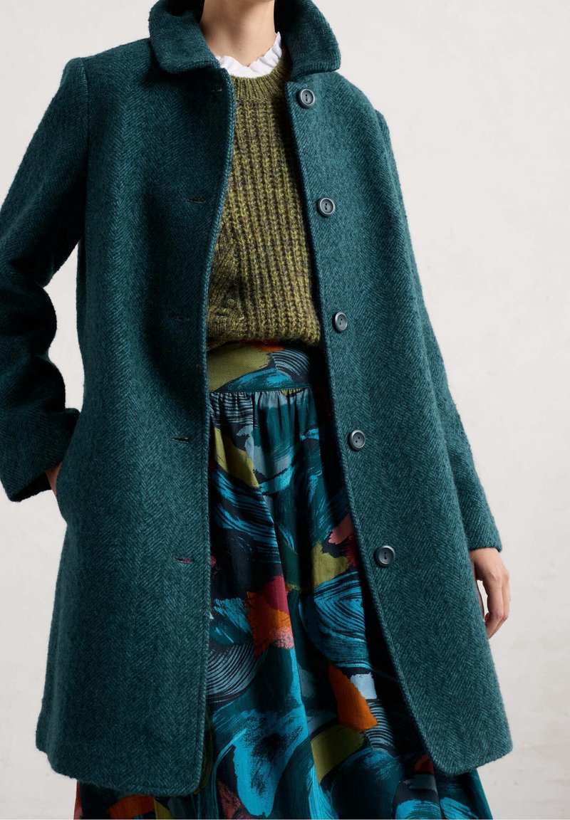 Donna che indossa un cappotto di lana blu verdi con bottoni, un maglione di lana verde oliva e una gonna colorata a motivi con un design a pennellate blu, arancioni e nere.