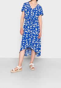 Vestido floral azul con escote en V, mangas cortas y un lazo en la cintura. El dobladillo es asimétrico, con un corte holgado y una tela ligera.