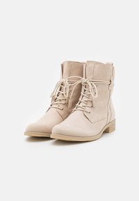Botas de tobillo de ante beige con cordones, tacones bajos y punteras redondas, exhibidas sobre un fondo blanco.