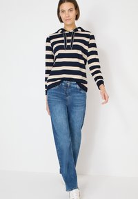 Marineblaue und cremefarbene gestreifte Kapuzenjacke mit Kordeln, kombiniert mit hellblauen Jeans mit geradem Bein und weißen Turnschuhen, vor einem schlichten Hintergrund.