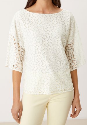 Vrouw draagt een witte kanten top met een bloemig bladmotief en ellebooglange mouwen, gecombineerd met licht crèmekleurige broek.