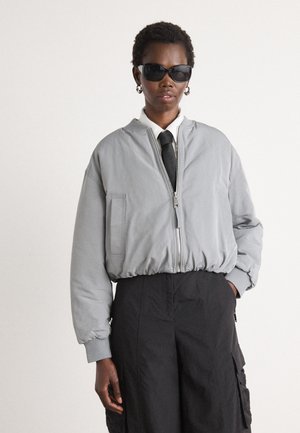 Grijze bomberjack met een ritssluiting, geribde manchetten en zoom. Gestyled met een wit shirt en een zwarte stropdas. Zwarte cargobroek.