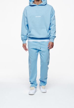 Sudadera con capucha azul claro con bolsillo frontal y logo blanco, combinada con pantalones deportivos a juego. Zapatillas blancas completan el look deportivo.