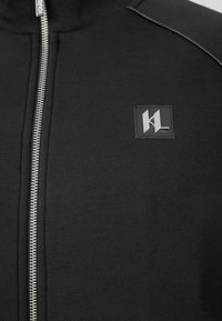 Giacca nera con zip e collo alto, caratterizzata da dettagli in argento, tessuto strutturato e una toppa con logo sul petto.