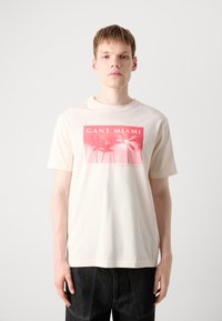 Krátký rukáv krémového trička s růžovým potiskem palem a textem "GANT MIAMI", vyrobeného z měkké bavlny.