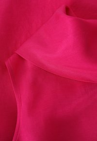 Felle roze stof met een gladde, glanzende textuur. Heeft een schuin kraag en een subtiele glans, die de zachtheid en de flow van het materiaal benadrukt.