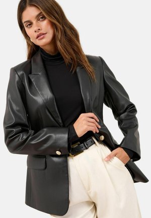 Black faux leather blazer with structured shoulders and gold button accents, paired with a black turtleneck and cream-colored trousers.