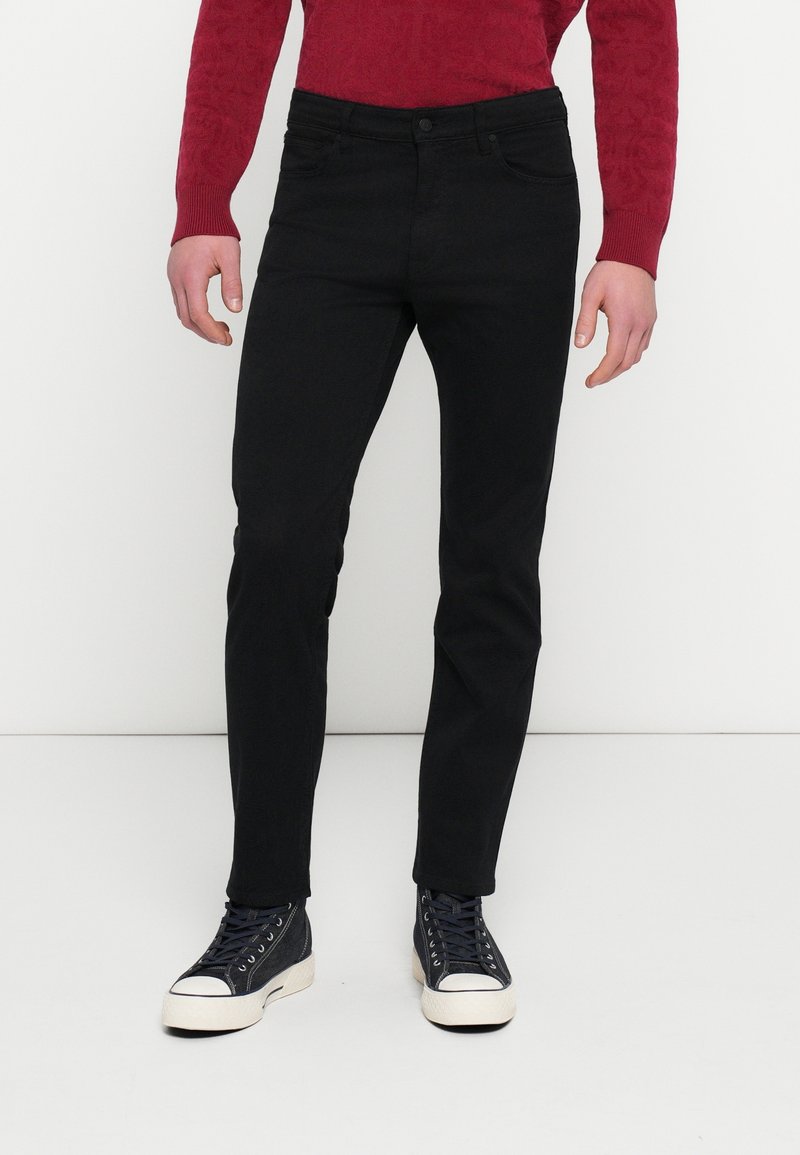Homme portant un jean slim noir, un pull rouge à manches longues et des baskets montantes noires avec semelles et bouts blancs.