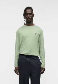 Μακρυμάνικη ανοιχτό πράσινη βαμβακερή μπλούζα με στρογγυλή λαιμόκοψη και μικρό μαύρο λογότυπο στο αριστερό μέρος του στήθους. Φορεμένη με σκούρο παντελόνι.