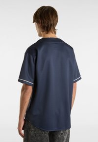 Donkerblauwe kortebloushirt van textuurstof, met witte afwerking op de mouwen en een ronde onderkant. De achterkant toont de kraag en stikdetails.