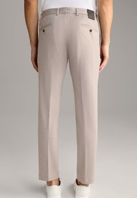 Ljusbeige byxor i en texturerad tygkvalitet, med två bakfickor med knäppning och en elegant, skräddarsydd passform. Vita sneakers synliga.