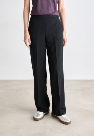 Frau trägt schwarze gerade geschnittene Hose, lila ärmelloses Oberteil und silber-weiße Sneakers, steht auf einem hellgrauen Boden vor einer schlichten Wand.