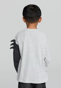 Maglietta grigia a maniche lunghe con maniche nere a contrasto, caratterizzata da un design di ritaglio unico sulla manica sinistra. Solo vista posteriore.