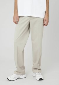 Un pantalon taillé beige clair avec une coupe décontractée, doté d'une texture lisse et de plis repassés, associé à des chaussures de sport blanches.