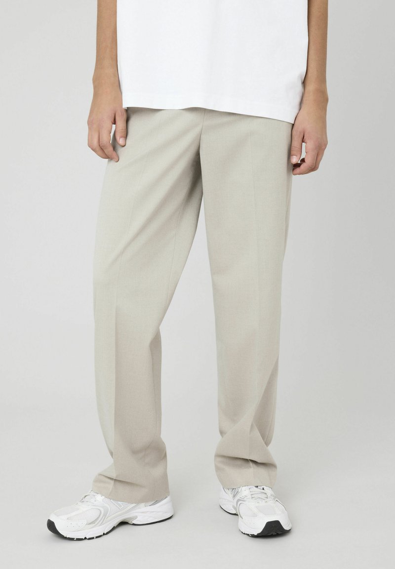Un pantalon taillé beige clair avec une coupe décontractée, doté d'une texture lisse et de plis repassés, associé à des chaussures de sport blanches.
