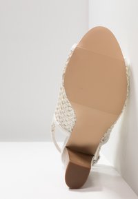 Bílé sandály na vysokém podpatku s textilním svrškem, hladkou béžovou podešví a štíhlým nártem. Spodek má rýhovaný vzor pro lepší přilnavost.