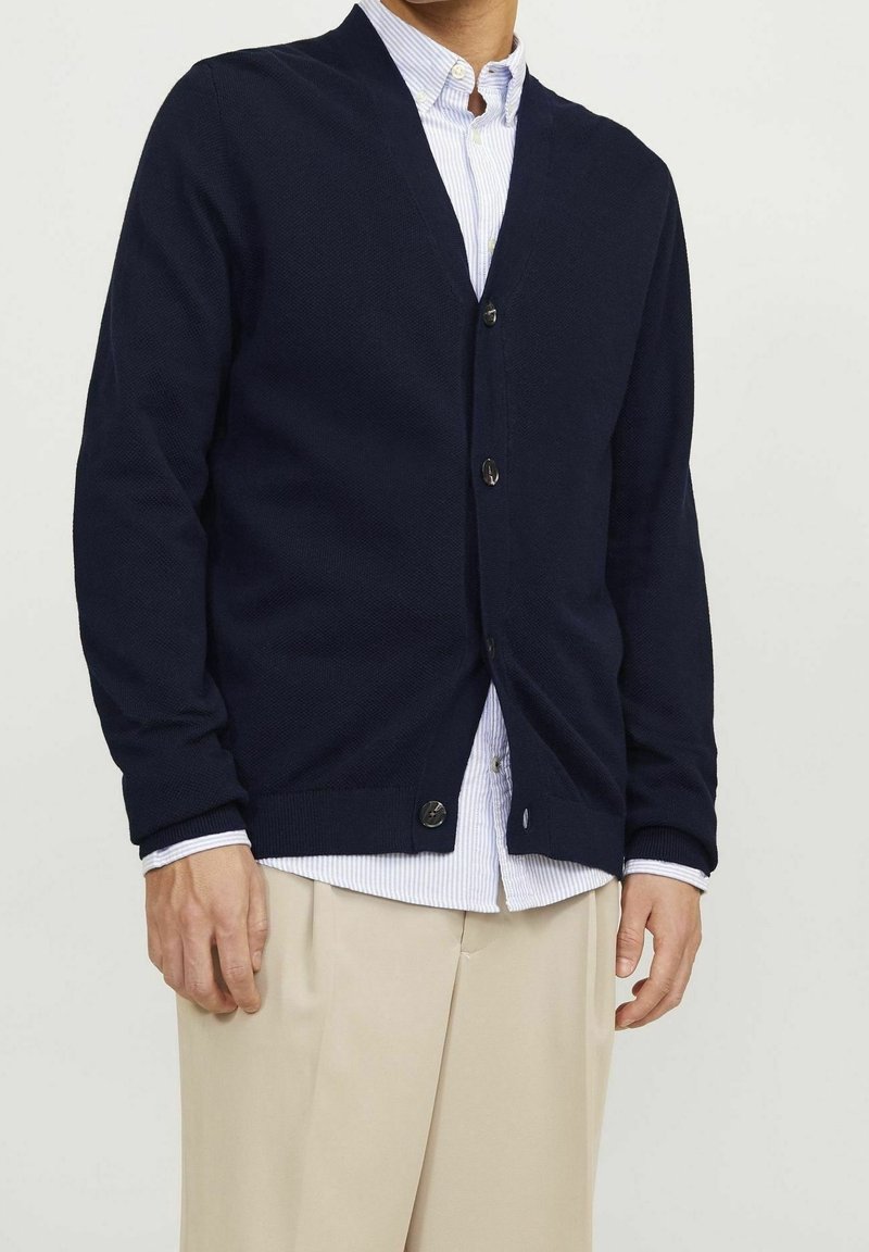 Marinblå V-ringad cardigan i texturerad stickad kvalitet, med långa ärmar, knäppning fram och ribbade cuffs, kombinerad med en randig skjorta.