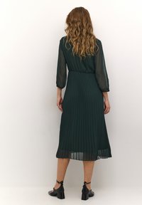 Mörkgrön plisserad midiklänning med genomskinliga ärmar och bältesdetalj, med en flödande design och texturerad tyg. Svarta fotledsskor med remmar.