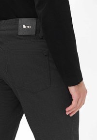 Jean en denim noir avec une finition texturée et des coutures visibles. Présente un patch rectangulaire en cuir portant l'inscription « Brax » sur la ceinture.