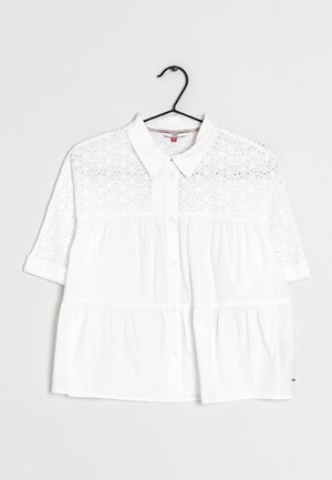 Blouse blanche à manches courtes boutonnée avec col, motif œillet sur la partie supérieure et les manches, suspendue à un cintre noir sur fond blanc.