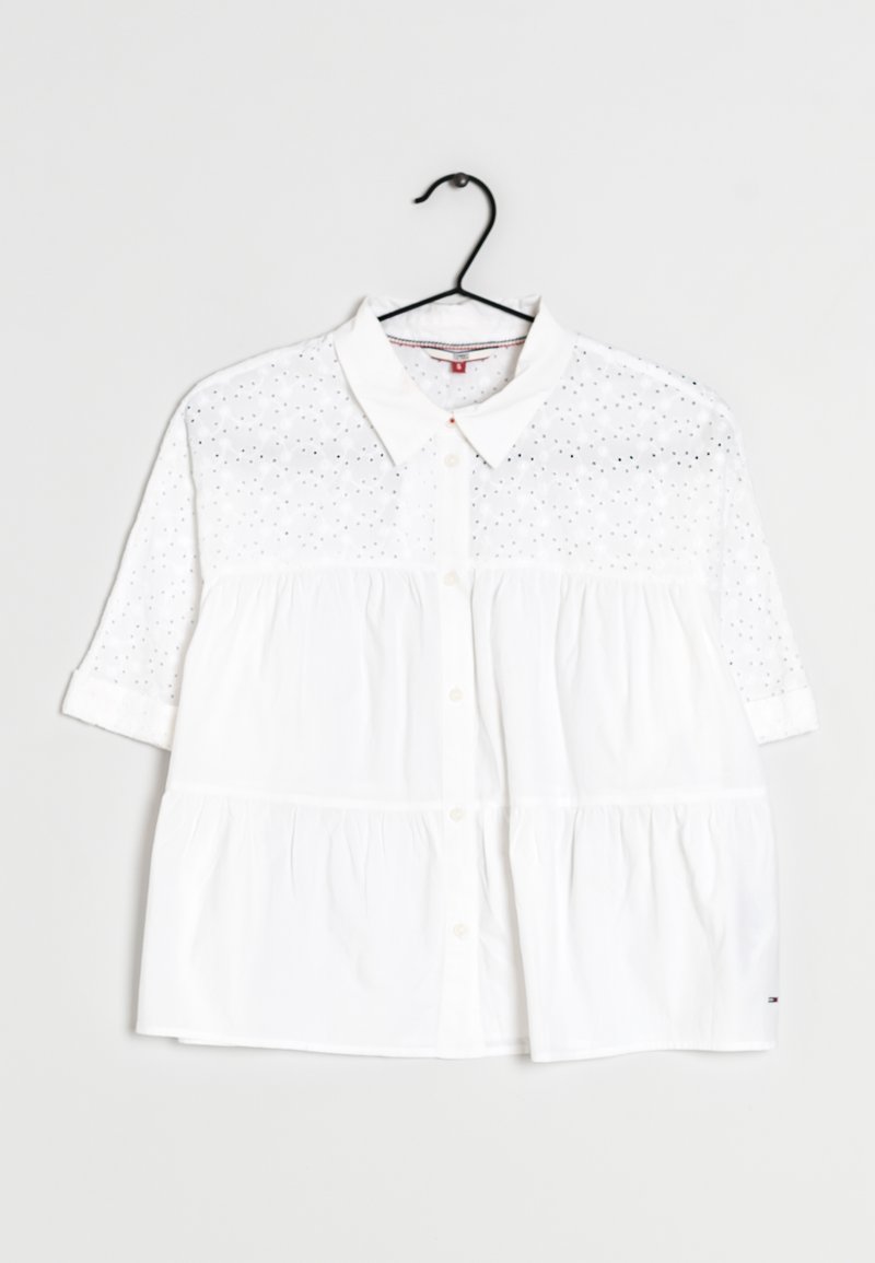 Blouse blanche à manches courtes boutonnée avec col, motif œillet sur la partie supérieure et les manches, suspendue à un cintre noir sur fond blanc.