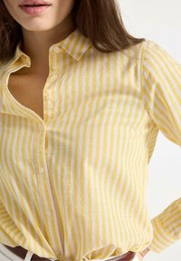Gul- og hvidstribet langærmet skjorte med krave, der har knapper og en let stofstruktur. Nærbillede af halsudskæringen.
