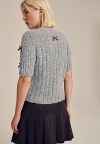 Pull gris clair tricoté avec une texture côtelée et des manches courtes, avec des nœuds à carreaux rouges sur les épaules. Associé à une jupe foncée.
