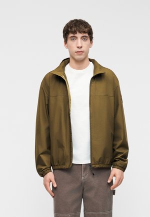 Mladý muž oblečený v olivovo zelenej bunde na zips cez bielu košeľu a sivé nohavice, stojaci pred jednoduchým svetlosivým pozadím.