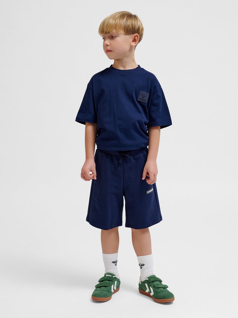 Młody chłopiec patrzy na bok, ubrany w granatowy t-shirt i krótkie spodenki, białe skarpetki oraz zielone trampki, stoi na jednolitym tle.