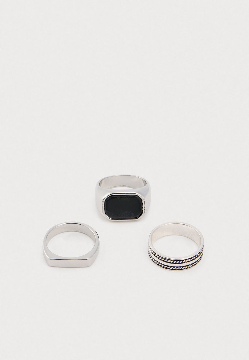 Tres anillos de plata sobre una superficie blanca: uno con una piedra rectangular negra, uno liso y uno con detalles de bandas que parecen cuerda negra.