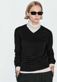Maglione nero a V con un dolcevita crema sotto. Il maglione ha maniche lunghe e polsini a costine. Modello con occhiali da sole scuri e rettangolari.