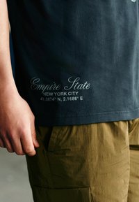 Tumši zilas krāsas kokvilnas T-krekls ar baltu tekstu "Empire State" un koordinātēm. Valkāts ar khaki kravas šortiem, kuros redzama kabatu detaļa.