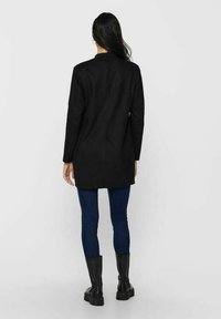 Cappotto lungo nero con un design su misura, caratterizzato da maniche lunghe e una cucitura posteriore pulita, abbinato a jeans blu scuro e stivali neri alla caviglia.