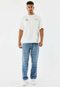 Joven de pie con camiseta gráfica blanca, jeans azules con texto repetido "Project X Paris" y zapatillas blancas sobre fondo blanco.