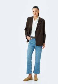 Blazer marrón de corte cruzado sobre una camisa blanca, combinado con jeans acampanados de un azul claro y mocasines beige. Texturas suaves y diseño clásico.