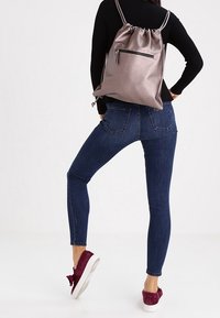 Šedý batoh na stažení vyrobený z hladkého materiálu, s přední kapsou na zip. Nosený s tmavými slim džíny a burgundskými teniskami.