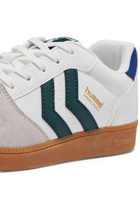 Hvid sneaker med grønne og sorte striber, brun gummisål og detaljer i ruskind. Har "hummel"-logo i guld. Snørebåndene er hvide.